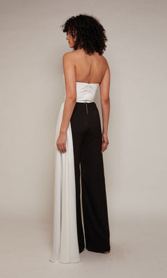 A person stands facing away, wearing the Long Formal Jumpsuit: Alyce 70050—an elegant strapless style with a white top, black pants, and a draped white panel on one leg—set against a plain light background.