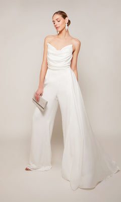 A woman wears the Alyce 70050 long formal jumpsuit featuring a strapless satin design with a draped bodice and wide legs, accessorized with a silver clutch and statement earrings.
