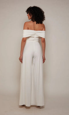 A person with curly hair stands facing away, wearing the Alyce 70051 long white off-the-shoulder satin jumpsuit with wide-leg pants against a plain light background.