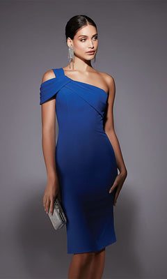 A woman stands against a plain gray background wearing the Alyce 70054 knee-length semi-formal dress in blue, styled with a silver clutch, long earrings, and a sleek updo.