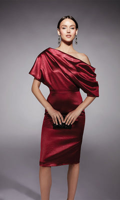 Woman in a wine-colored, off-shoulder Alyce 70060 knee length semi-formal dress holds a small black clutch, standing against a plain grey background.
