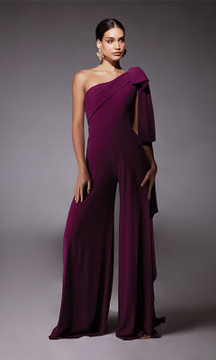 A person wearing the Alyce 70065 long formal jumpsuit in deep purple, featuring one-shoulder styling, wide legs, and a large bow detail—an elegant cocktail dress alternative—stands against a plain gray background.