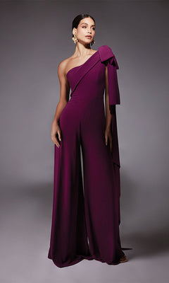 A woman wears the Alyce 70065, a long formal purple one-shoulder jumpsuit with a large bow detail and wide-leg pants, paired with gold statement earrings. She stands against a grey background—a chic alternative to a cocktail dress.