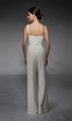 A woman faces away, wearing the Alyce 70073 Long Formal Jumpsuit—featuring a sleeveless white lace top with floral embroidery and wide-legged pants—set against a plain gray background.