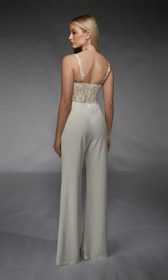 A woman faces away in the Alyce 70074 Long Formal Jumpsuit, featuring a white design with lace and floral embroidery on the back and wide-leg pants, set against a plain gray background.