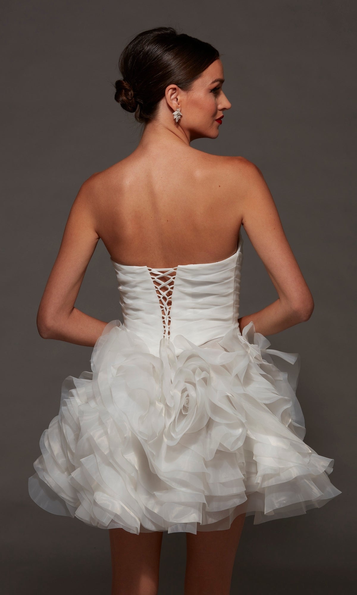 A woman wearing the Short White Party Dress: Alyce 70086, featuring a strapless sweetheart neckline, lace-up corset back, and layered ruffled skirt, stands with her hands on her hips against a plain background.