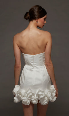 A woman faces away, wearing the Alyce 70087 short white party dress featuring a strapless bodycon silhouette with a fitted bodice and large rosette hemline details, set against a plain dark background.