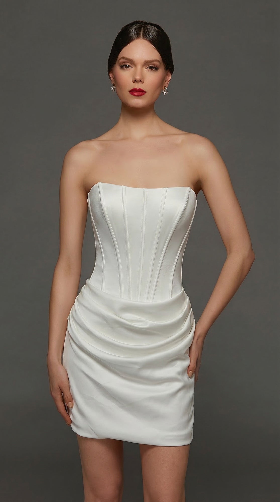 A woman wears the Alyce 70090 Short White Party Dress in Diamond White, featuring a strapless satin design with a structured corset bodice, as she stands against a plain dark background with her hands relaxed at her sides.