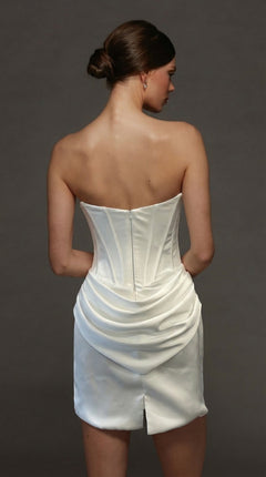 A woman with dark hair in a low bun is seen from the back against a plain dark background, wearing the Alyce 70090 short white party dress featuring a strapless satin design with a fitted corset bodice and draped skirt.