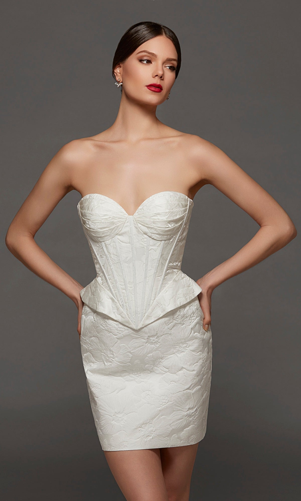 The model wears the Alyce 70093 Short White Party Dress in Diamond White, featuring a strapless sweetheart neckline, a structured corset bodice, and a textured white brocade finish as she poses with hands on hips against a plain gray background.