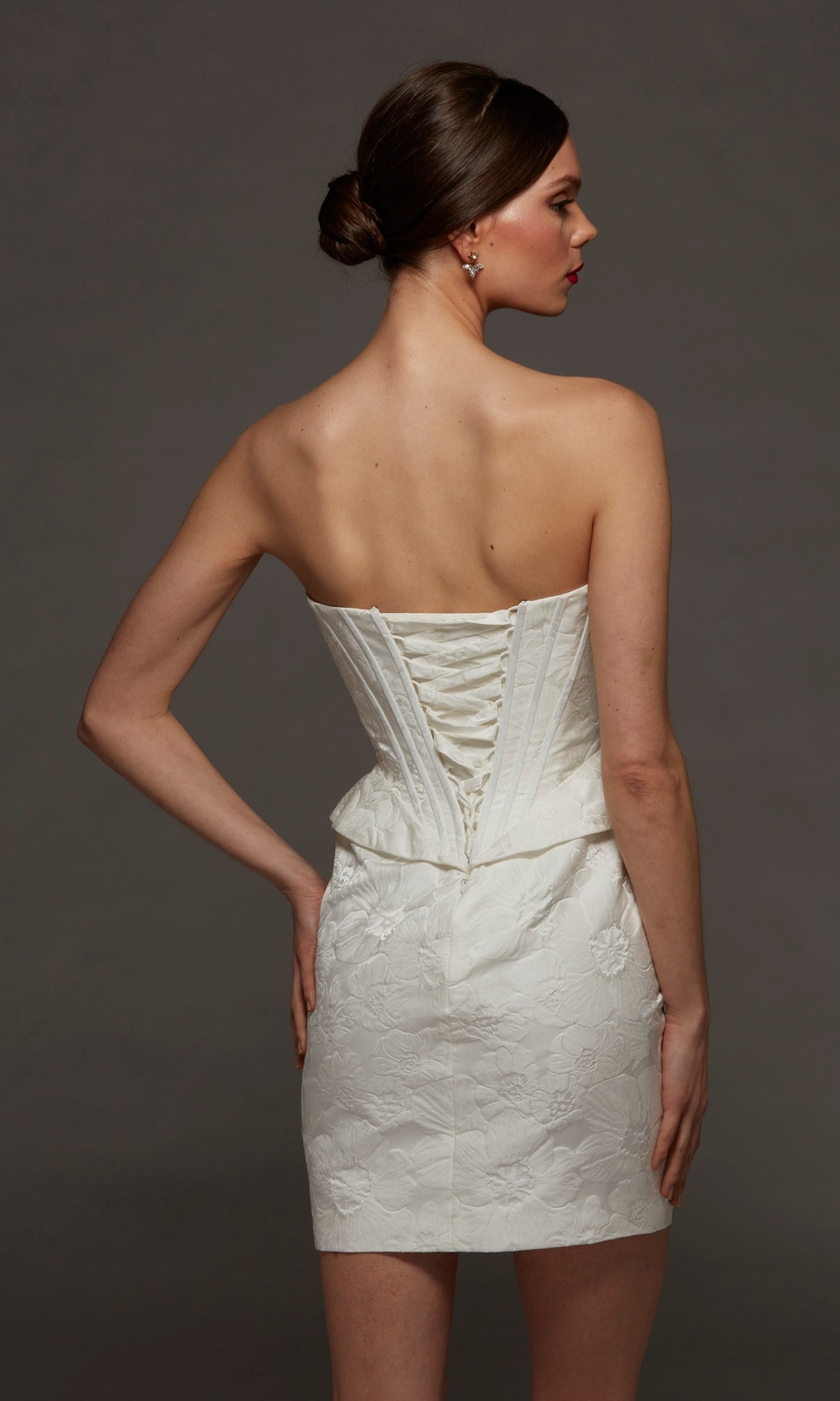 A woman stands against a plain dark background wearing the Alyce 70093 Short White Party Dress, a strapless mini dress with a sweetheart neckline, lace-up back, delicate floral pattern, and corset bodice for a flattering fit.