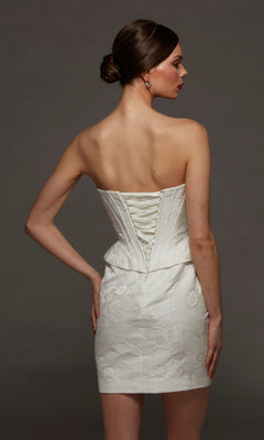 A woman stands against a plain dark background wearing the Alyce 70093 Short White Party Dress, a strapless mini dress with a sweetheart neckline, lace-up back, delicate floral pattern, and corset bodice for a flattering fit.