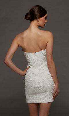 A woman stands with her back to the camera against a plain dark background, wearing the Alyce 70095 short white party dress—a strapless, fitted jacquard mini dress featuring a corset bodice.