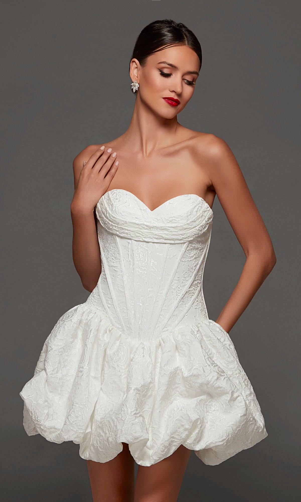 A woman poses against a plain gray background wearing the Alyce 70096 short white party dress in diamond white, featuring a strapless jacquard design with a fitted corset bodice and a voluminous skirt.
