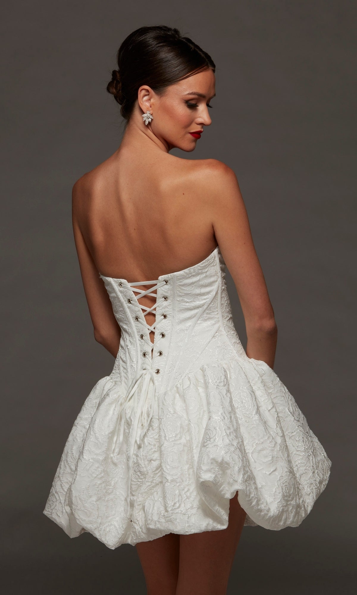 A woman wearing the Alyce 70096 short white party dress, featuring a strapless sweetheart neckline, jacquard textured fabric, lace-up back, and a voluminous short skirt, stands against a plain dark background.