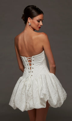 A woman wearing the Alyce 70096 short white party dress, featuring a strapless sweetheart neckline, jacquard textured fabric, lace-up back, and a voluminous short skirt, stands against a plain dark background.