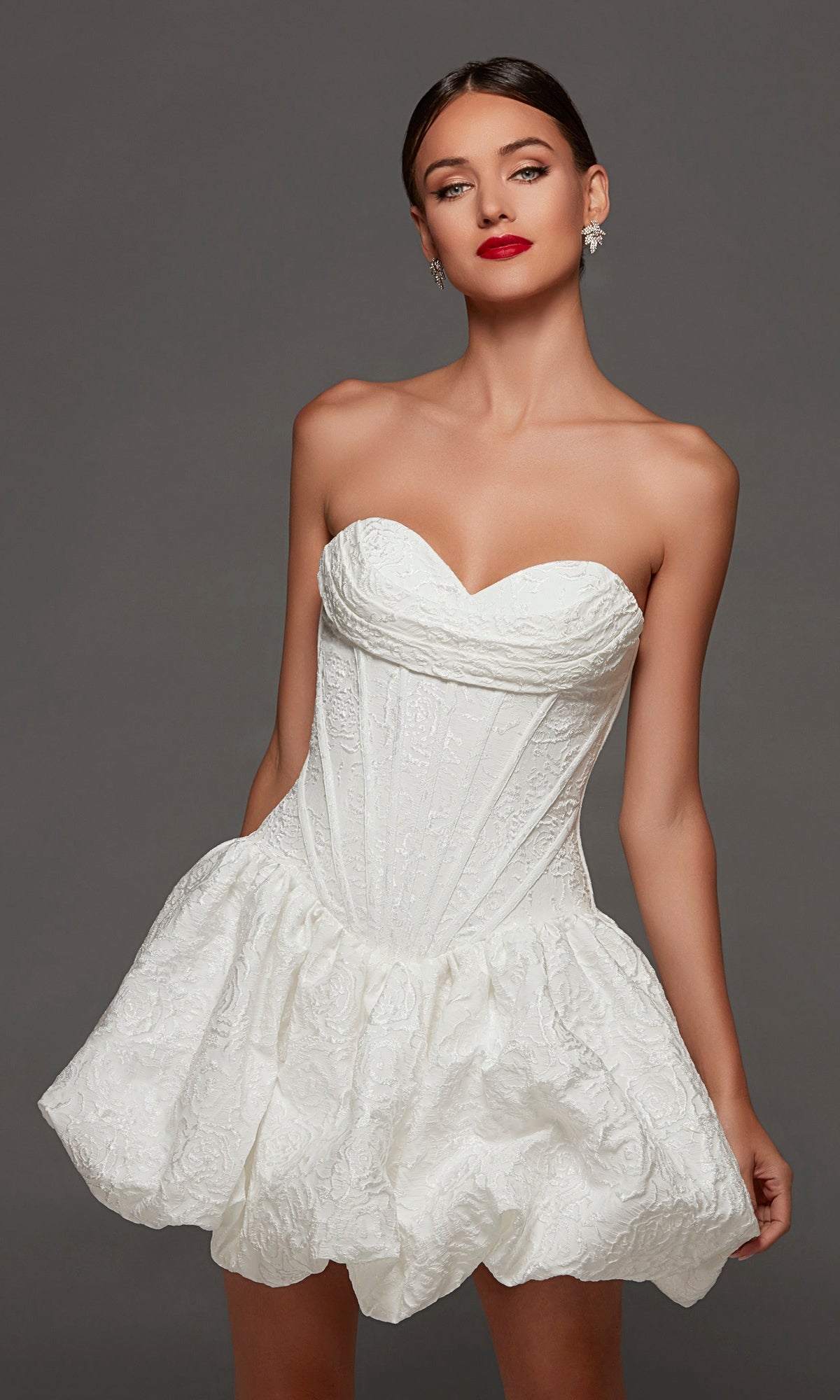A woman models the Alyce 70096 short white party dress, which features a strapless jacquard design with a fitted corset bodice and flared skirt, posing against a plain gray background.