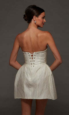 A woman is wearing the Alyce 70097 short white party dress, a strapless jacquard mini dress with a textured finish and lace-up back detail. Standing against a dark background with her hands on her hips, she showcases this perfect short party dress for any occasion.