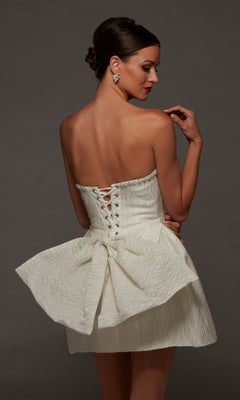 Woman wearing the Alyce 70097 short white party dress—strapless with jacquard fabric and a large bow at the back—posing against a plain dark background.