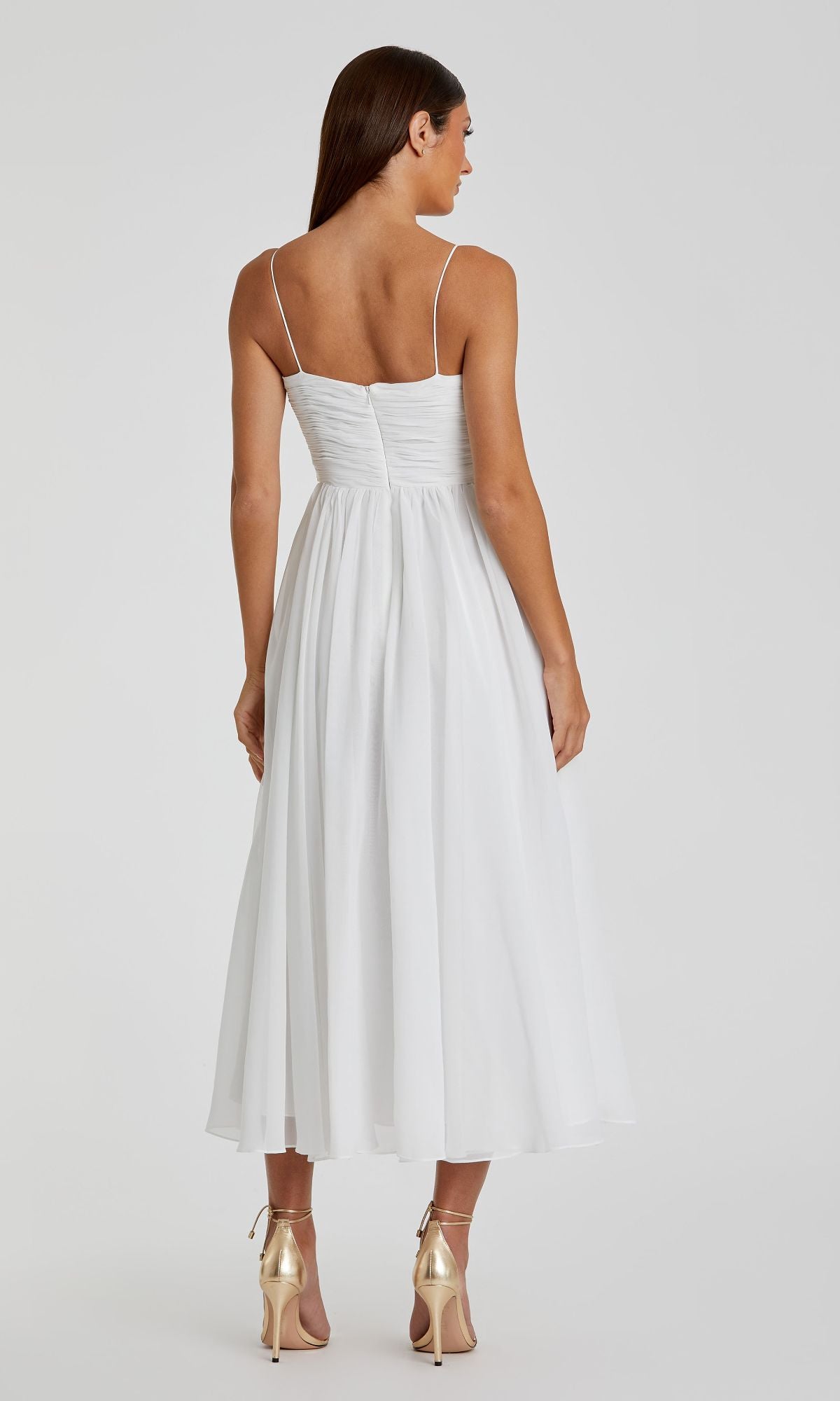A woman in the Mac Duggal 70099 A-Line Midi Wedding Guest Dress—white, sleeveless, tea-length with thin straps—and gold high heels stands with her back to the camera.