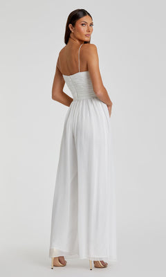 A woman poses backward, looking over her shoulder, in the Mac Duggal 70100 Flowing Wide-Leg Formal Jumpsuit. The white, sleeveless jumpsuit features thin straps and wide-leg pants.