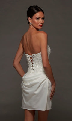 Model wearing the Alyce 70105 short white party dress, featuring a strapless corset bodice with lace-up back, beaded detailing, and a satin skirt, posed against a plain dark background.