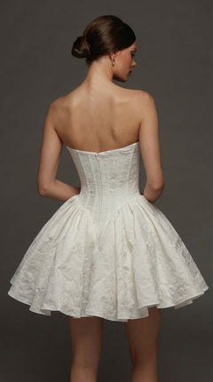 A woman in the Alyce 70110 Short White Party Dress, featuring a strapless, textured fit-and-flare silhouette with a sweetheart neckline, stands against a plain dark background facing away from the camera.