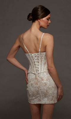 A woman stands with her back to the camera against a plain dark background, wearing the Alyce 70115 short white party dress featuring delicate lace and a lace-up back.