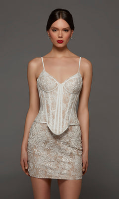 A woman wearing the Alyce 70115 Short White Party Dress, featuring a sleeveless, sequined lace design with a structured corset bodice and delicate lace-up back, stands against a plain gray background.