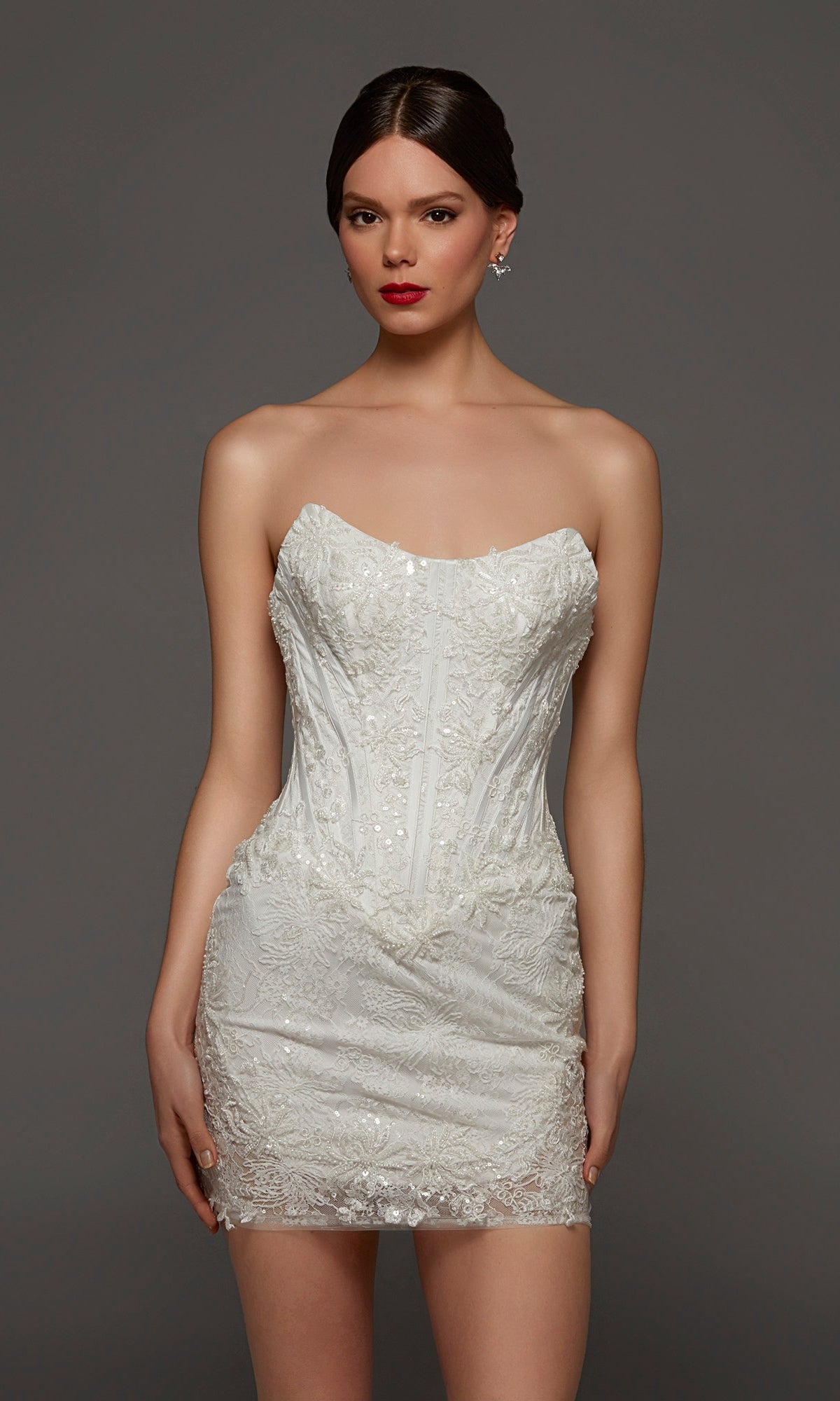 A woman wearing the Alyce 70116 Short White Party Dress in Diamond White, featuring a strapless, fitted silhouette with delicate lace detailing and a flattering corset bodice, stands against a plain dark background.