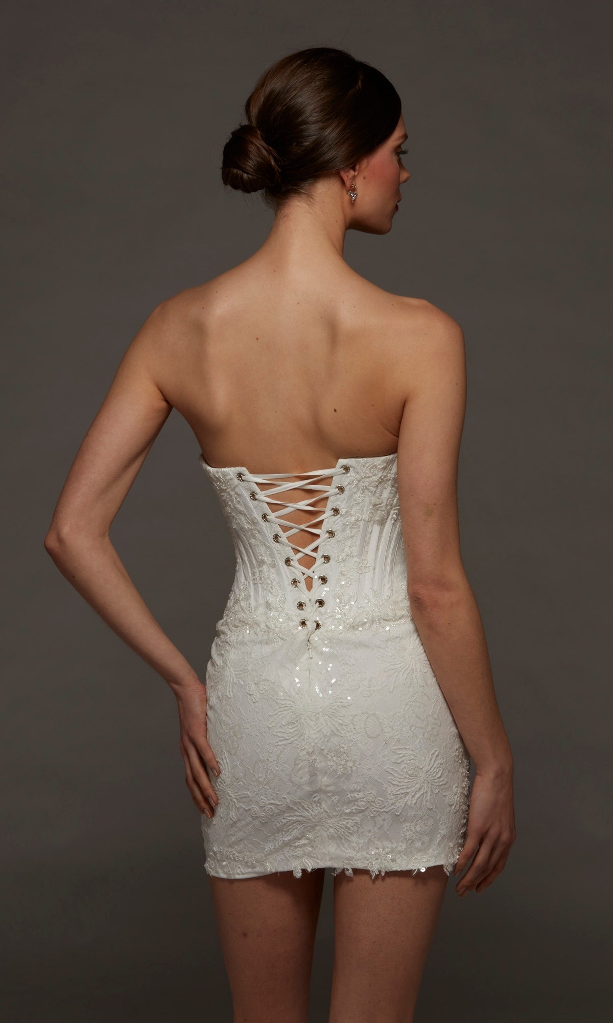 A woman faces away, wearing the Alyce 70116 short white party dress, which features a strapless scoop neckline and an elegant corset bodice with lace-up detailing at the back. Her hair is styled in a neat bun as she stands against a plain dark background.