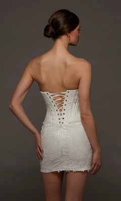 A woman faces away, wearing the Alyce 70116 short white party dress, which features a strapless scoop neckline and an elegant corset bodice with lace-up detailing at the back. Her hair is styled in a neat bun as she stands against a plain dark background.