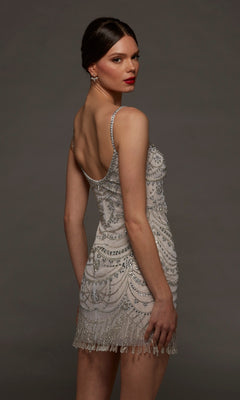 Model showcases the Alyce 70123 short party dress— a sleeveless, bead-embellished silver style featuring a beaded fringe hem—while posing against a dark background and looking over her shoulder with a neutral expression.