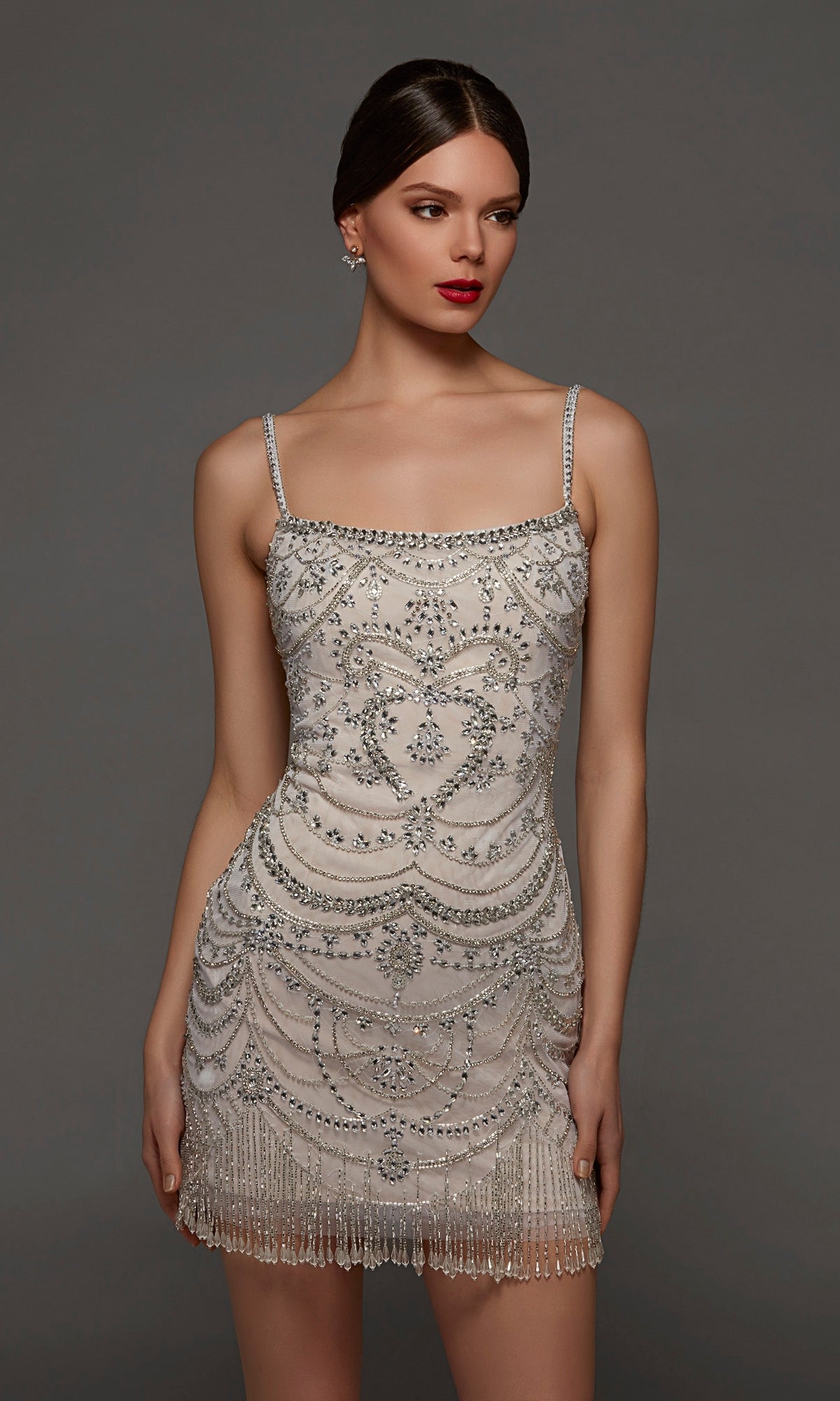 A woman stands against a plain gray background wearing the Short Party Dress: Alyce 70123, a sleeveless mini dress adorned with intricate silver embellishments and shimmering beaded fringe at the hem. This chic party dress exudes modern elegance with its dazzling details.