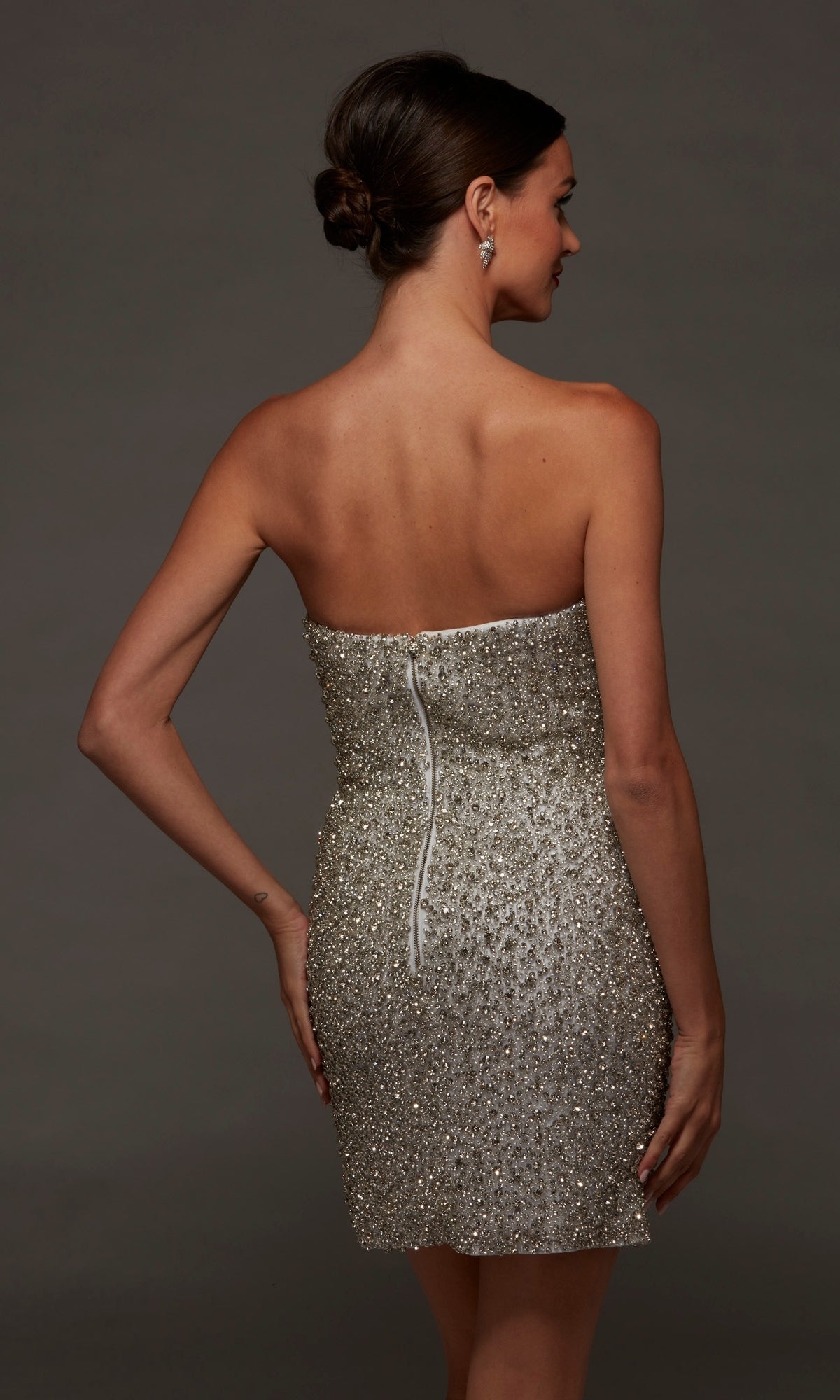 A woman wearing the Short White Party Dress: Alyce 70124, featuring a strapless beaded design and a fitted silhouette, stands with her back to the camera and hand on her hip against a plain dark background.