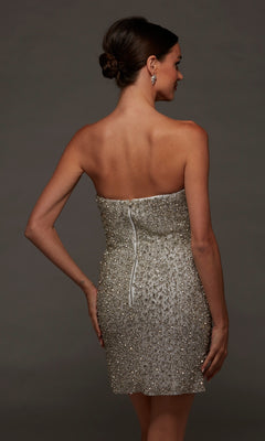 A woman wearing the Short White Party Dress: Alyce 70124, featuring a strapless beaded design and a fitted silhouette, stands with her back to the camera and hand on her hip against a plain dark background.