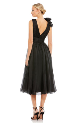 A woman wears the Mac Duggal Short Wedding Guest Dress 70165, a black knee-length dress with a deep V-back, ruched ruffled bodice, and one-shoulder bow detail, paired with black high-heeled sandals as she stands facing away.
