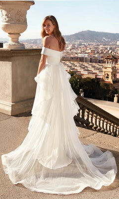 A woman wears the Alyce 7020 long white dress on a terrace, cityscape and hills behind her, highlighting the gown's elegant bridal style.
