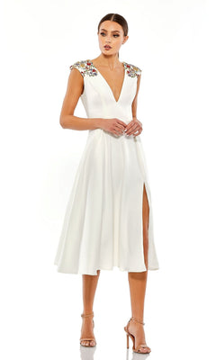 A woman wears the Short Wedding Guest Dress 70214 by Mac Duggal, a white beaded knee-length party dress with a deep V-neck, floral shoulder embellishments, front slit, and nude high heels.