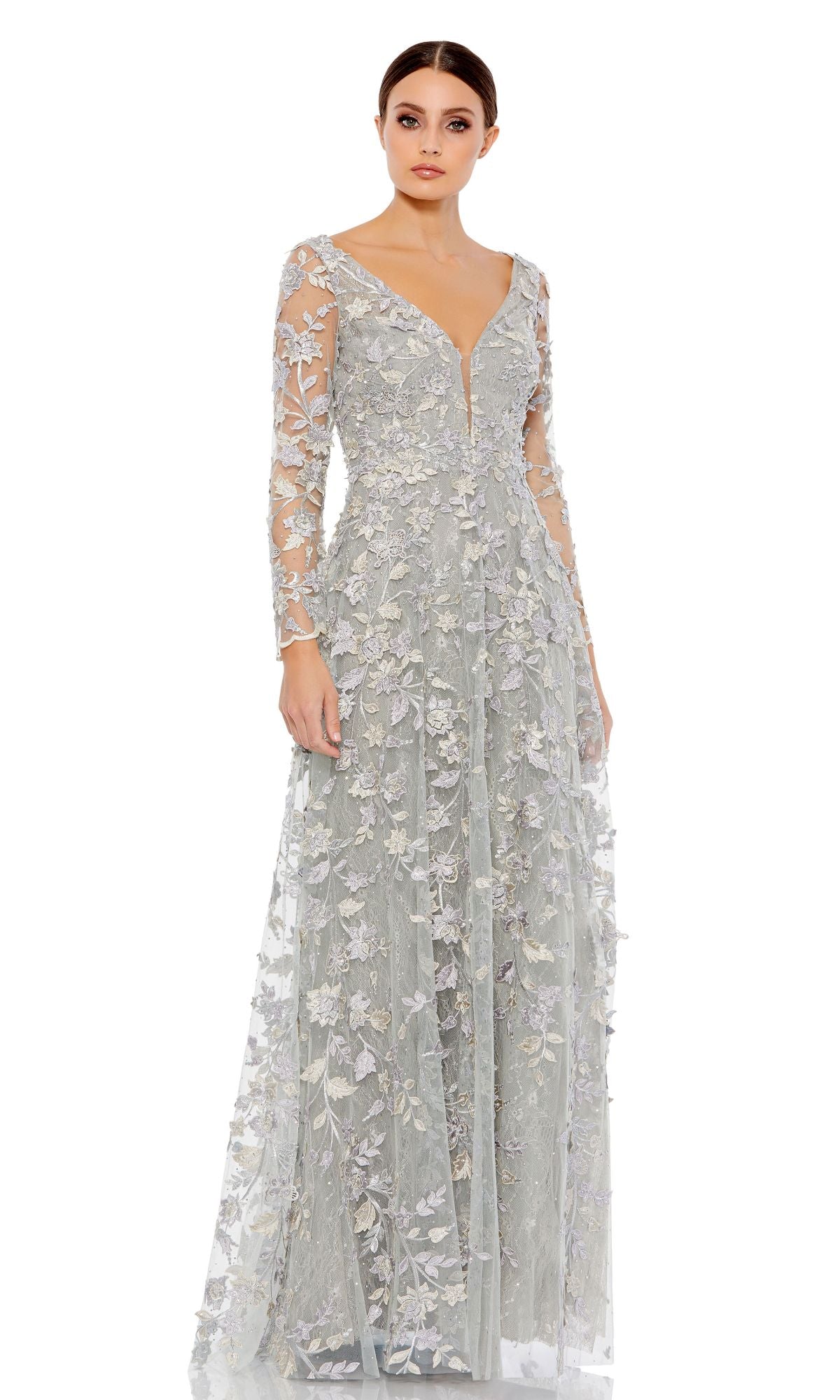 A woman wears the Mac Duggal 70224 Long Sleeve Long Formal Dress, a light gray embroidered maxi with sheer sleeves and floral lace appliqué, standing against a plain white background.