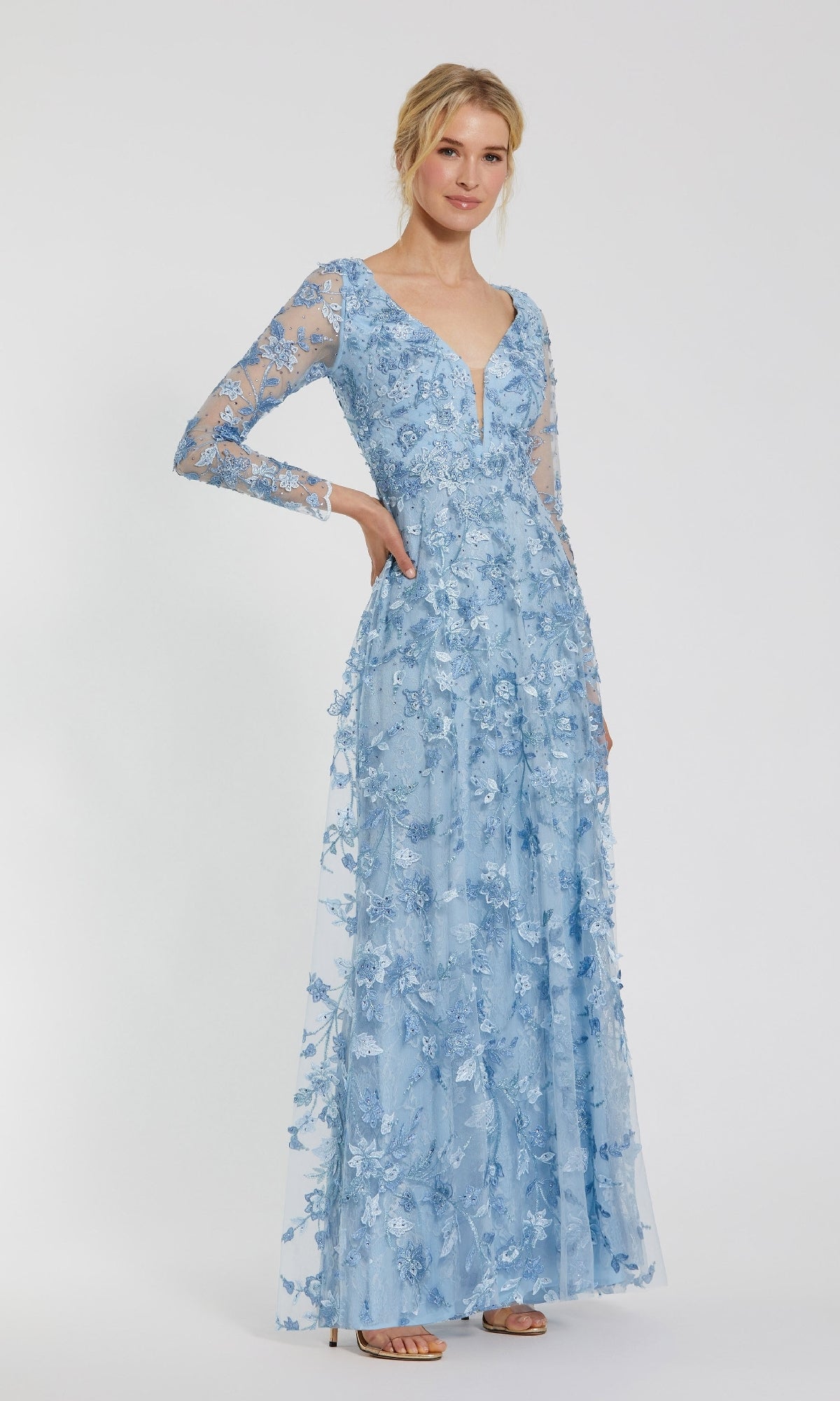 A woman models the Mac Duggal 70224 Long Sleeve Long Formal Dress, featuring light blue lace, floral embroidery, and sheer sleeves, standing against a plain white background.