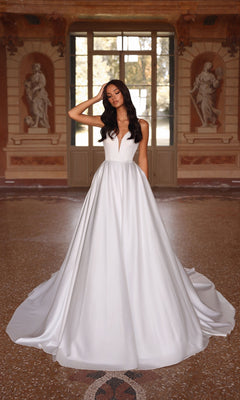 A woman wearing the Long Formal Dress: Alyce 7024 in diamond white, featuring a strapless bodice and elegant A-line skirt, stands in an ornate room with large windows and classical statues.