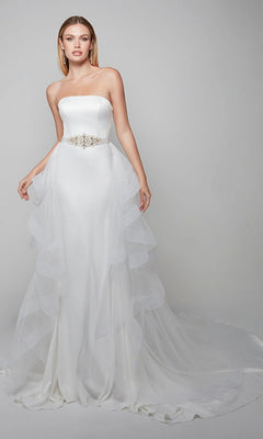 A woman stands against a plain background wearing the Alyce 7025, a long white strapless satin dress with a fitted bodice, jeweled belt, ruffle-layered tulle overskirt, and a long train.
