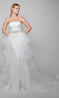 A woman wears the Alyce 7025 long white dress, a strapless formal gown with a fitted bodice, waist belt, and layered tulle overskirt with a long train, standing against a plain backdrop.