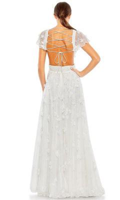 A woman with a low ponytail stands facing away, wearing the Mac Duggal Long Formal Dress 70275—an embellished, floral-detailed A-line gown featuring a lace-up open back.