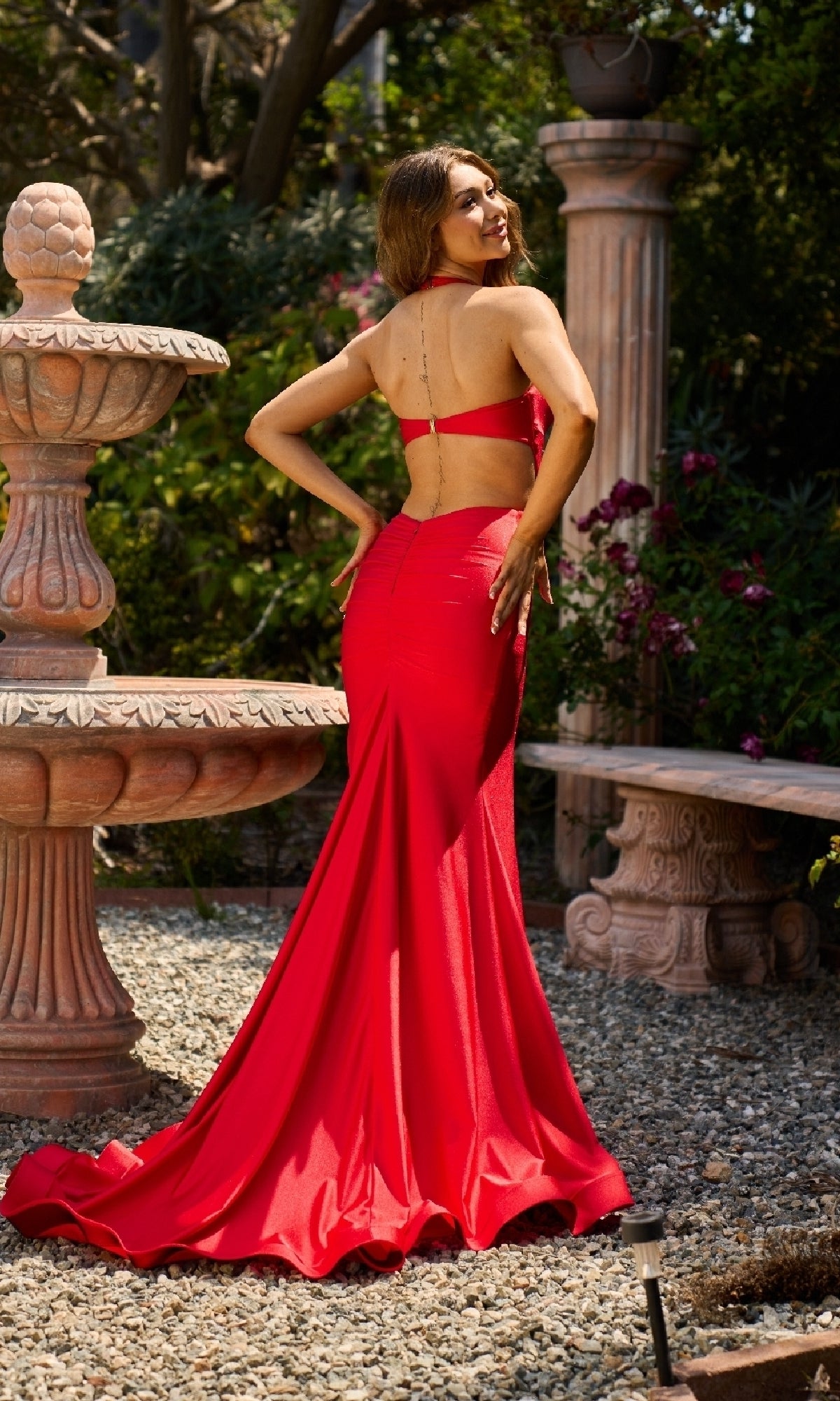 A woman wearing the Long Halter Prom Dress: Atria 7034H stands outdoors by a stone fountain and bench, looking over her shoulder with a smile.