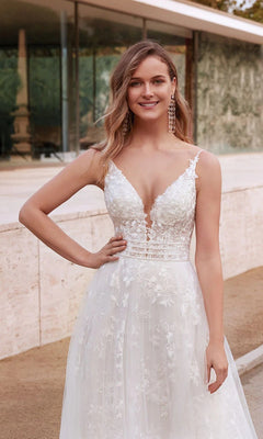 A woman stands outdoors, smiling at the camera in the Alyce 7036 Long White Dress with a deep V neckline and delicate floral appliqués.