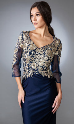 A woman stands against a gray background wearing the Amelia Couture 7039 Mother-of-the-Bride dress in navy, featuring an embroidered bodice and sheer three-quarter sleeves.