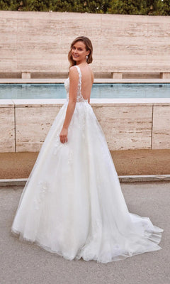 A woman in the Alyce 7039 long white dress, a sleeveless lace ball gown with a low back, stands outdoors and looks over her shoulder.