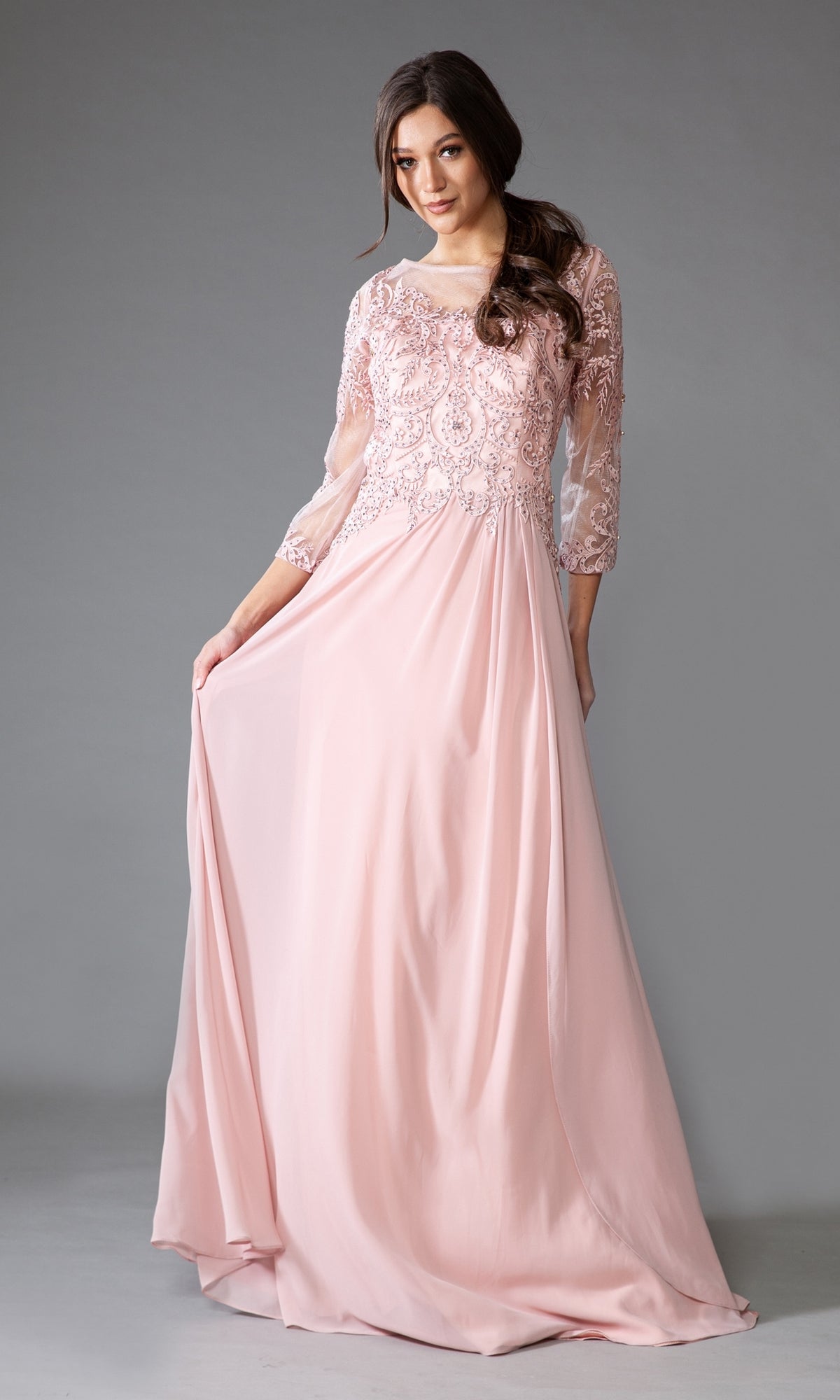 Woman modeling the Modest Long-Sleeve Mother-of-the-Bride Dress 7043 in light pink chiffon with lace bodice detailing and sheer three-quarter sleeves, standing against a gray background.